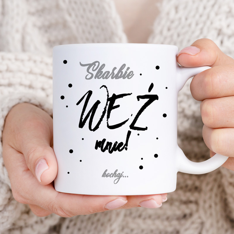 Biała filiżanka trzymana w dłoniach, zdobiona napisem "WEŹ mnie!" oraz dodatkowymi, ciemnymi kropkami.