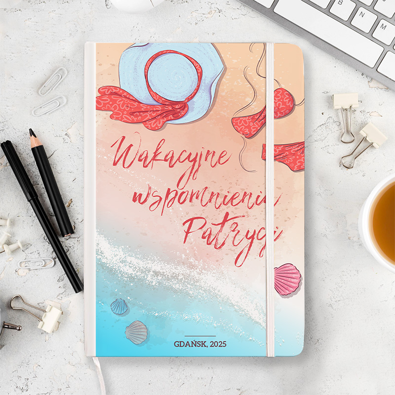 Notes z kolorową okładką przedstawiającą kapelusz, kostium bikini i muszle na plażowym tle, z napisem: "Wakacyjne wspomnienia Patrycji" i informacją, że jest to wydanie na rok 2025 z Gdańska, ustawiony na biurku obok długopisów i filiżanki z kawą.