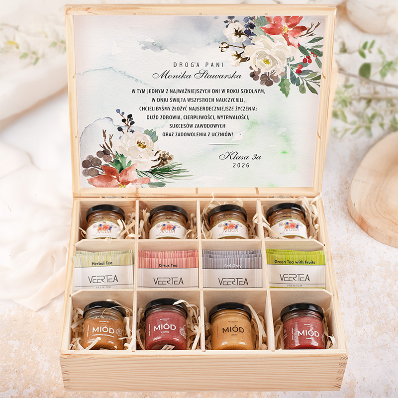 Drewniana skrzynka z prezentem, zawierająca słoiczki z miodem, herbatami oraz kartkę z życzeniami dla nauczycielki Moniki Stawarskiej. Na wieczku skrzynki znajdują się kwiatowe akwarelowe ilustracje oraz życzenia od klasy 3a na rok 2026.
