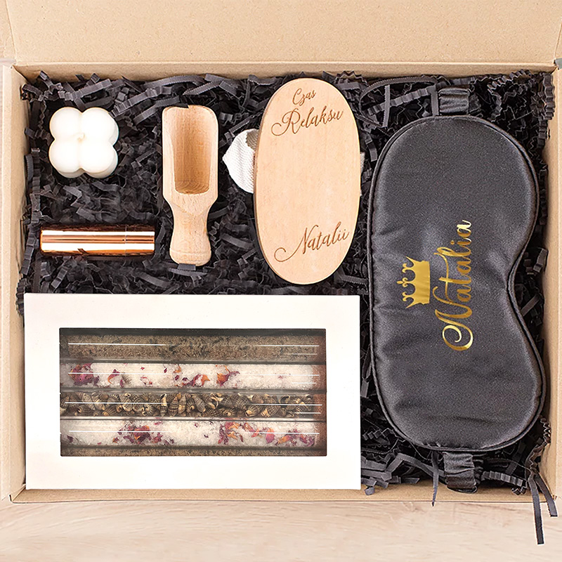 Zestaw prezentowy zawierający personalizowaną maskę do snu, drewnianą łyżkę, świeczki i słodkości w eleganckim opakowaniu.