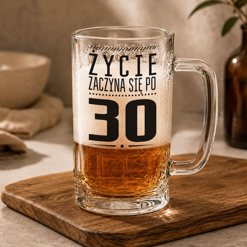 Szklanka piwa na drewnianej desce, z napisem "Życie zaczyna się po 30." w stylowej czcionce. W tle widać naczynie i butelkę.