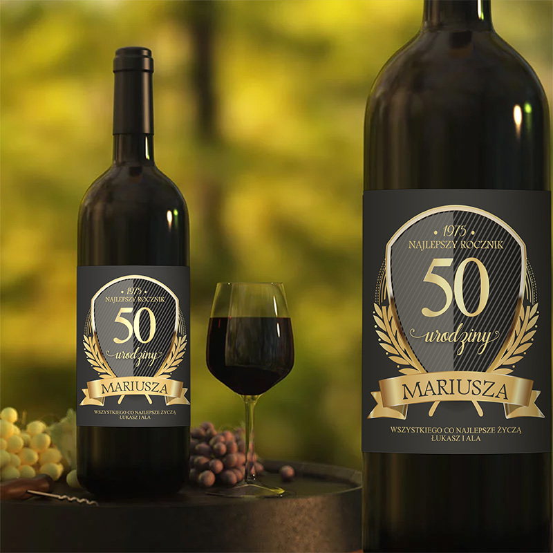 Butelka wina z etykietą z napisem "50 urodziny Mariusza", z datą 1975 i hasłem "Najlepszy rocznik". W pobliżu stoi kieliszek z czerwonym winem oraz winogrona na tle rozmytej zielonej scenerii.