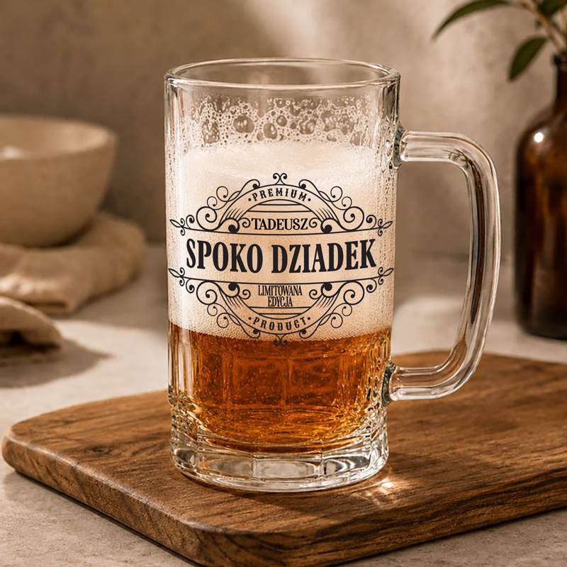 Szkło z piwem na desce, z napisem "Spoko Dziadek" i dekoracyjnymi detalami. W tle widać naczynia i butelkę.