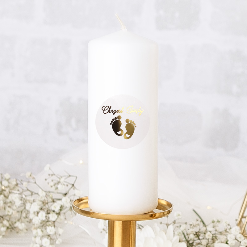 Świeca pieńkowa na Chrzest w białym kolorze. Do niej znajduje się etykieta ze złoconym nadrukiem Chrzest Święty i motyw stópek.