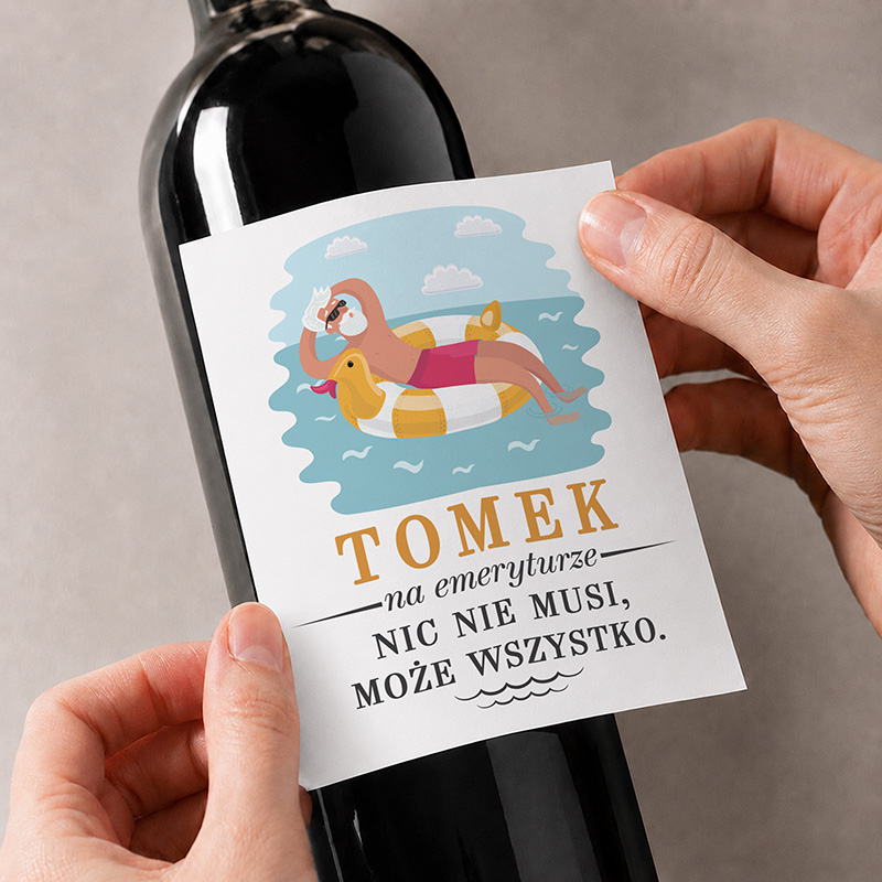 Etykieta na butelce wina z grafiką przedstawiającą osobę na dmuchanym materacu, tekst "TOMEK na emeryturze NIC NIE MUSI, MOŻE WSZYSTKO."