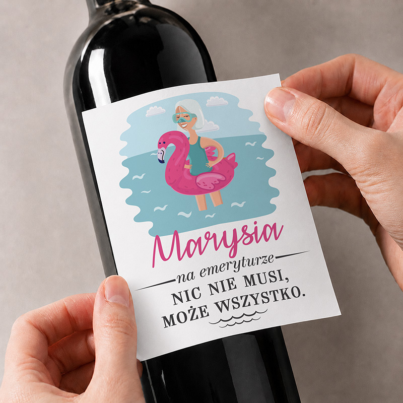 Butelka wina z etykietą przedstawiającą kobietę na różowym flamingu w wodzie z napisem "Marysia na emeryturze NIC NIE MUSI, MOŻE WSZYSTKO."