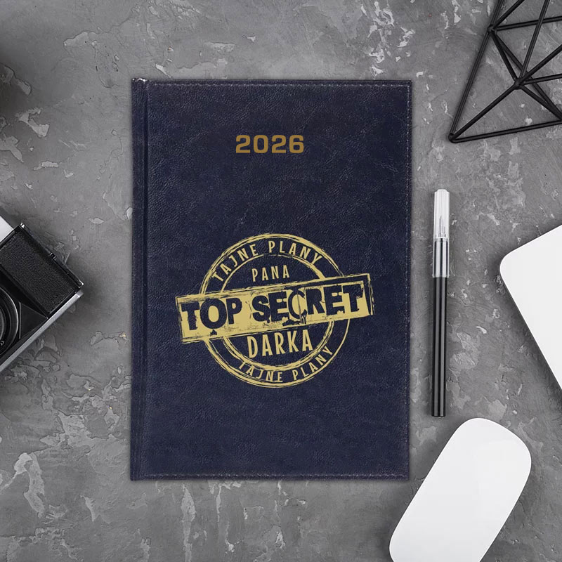 Ciemnoniebieski notes z napisem 'TOP SECRET' oraz rokiem 2026, leżący na szarej powierzchni, obok czarnego długopisu, białej myszki komputerowej i aparatu fotograficznego.