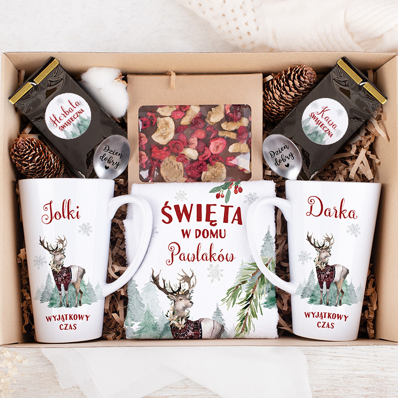 Zestaw prezentowy w kartonie zawierający dwie kubki z reniferami, ozdobny ręcznik z napisem "Święta w domu Pawlaków", kawę i herbatę, a także liście i kwiaty w ozdobnym opakowaniu, wszystko otoczone naturalnymi podkładkami.