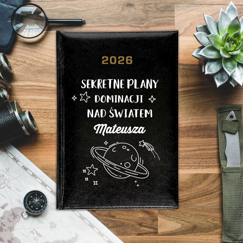 Notatnik z czarną okładką, na której widnieje napis "2026 SEKRETNE PLANY DOMINACJI NAD ŚWIATEM Mateusza" oraz rysunki planet i gwiazdek. W tle widoczne różne akcesoria, takie jak kompas, lupa, kamera i roślinka.