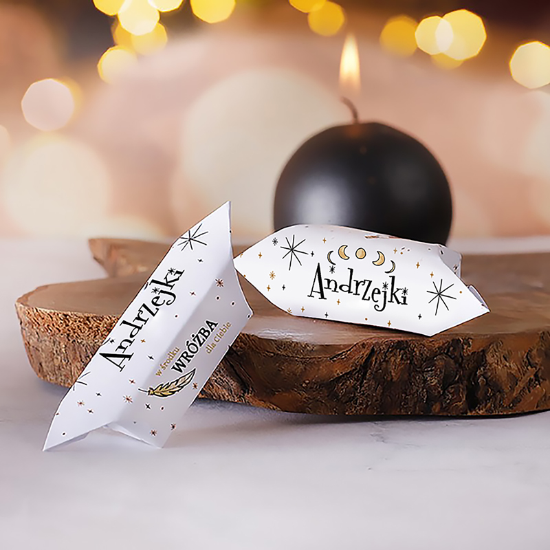 Dwa ciasteczka andrzejkowe o białych opakowaniach z czarnymi i złotymi napisami, leżące na drewnianym stole obok czarnej świecy, w tle delikatne, rozmyte światła.