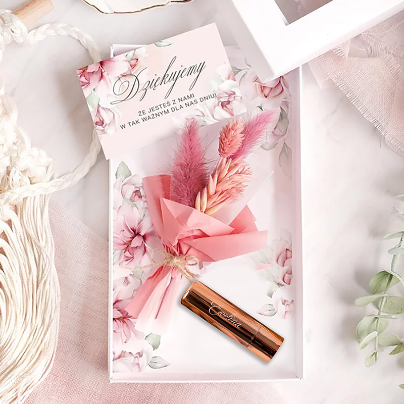 Pudełko z pastelowym tłem zawierające różowe kwiaty i suszone trawy, z umieszczonym na wierzchu różowym kosmetykiem w metalowej tubce.