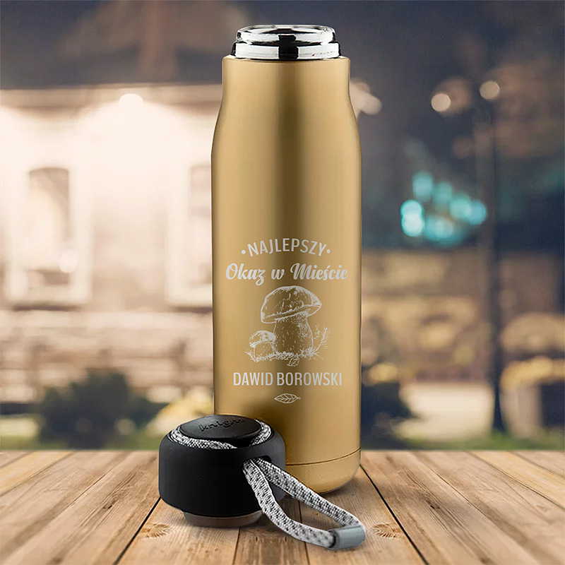Ozdobny termos w kolorze złotym z napisem 'Najlepszy Okaz w Mieście' oraz imieniem 'Dawid Borowski', obok czarna nakrętka z szarym sznurkiem, tło lekko rozmyte z widocznymi zarysem budynków i latarniami.