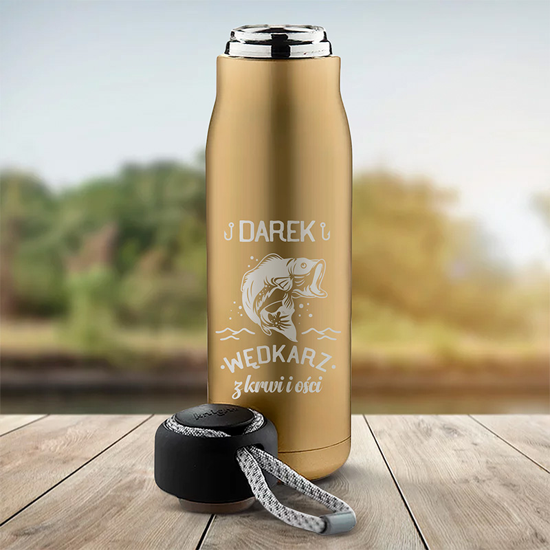 Metalowy termos w kolorze złotym z imieniem 'Darek' oraz napisem 'WĘDKARZ z krwi i kości', obok czarny uchwyt z szarym sznurkiem, w tle rozmyty krajobraz natury.