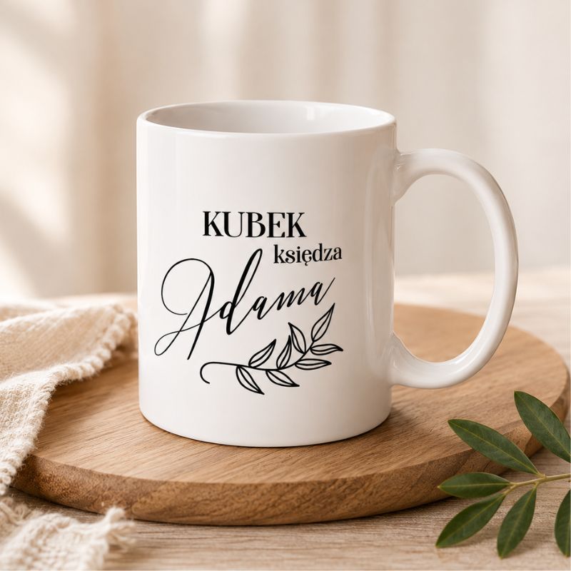 Biały kubek z napisem "KUBEK księdza Adama" oraz ozdobnym wzorem liści. Na drewnianej podstawie obok materiałowa serweta.