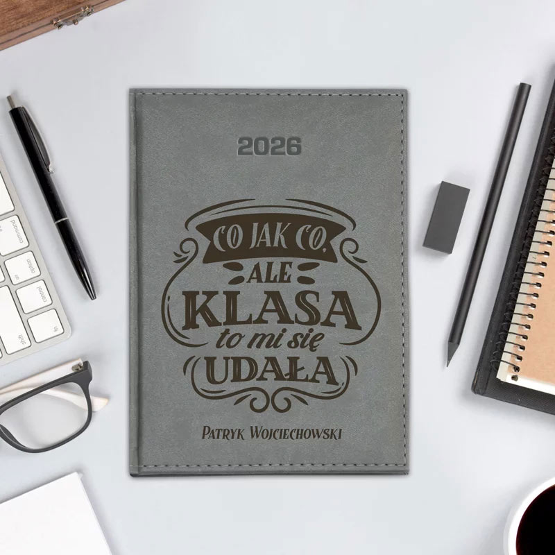 Szary notes z napisem "CO JAK CO, ALE KLASA TO MI SIĘ UDAŁA" oraz imieniem "Patryk Wojciechowski" i rokiem 2026, leżący na biurku obok długopisu, okularów, ołówka, gumki do mazania, klawiatury komputerowej oraz notatnika ze spiralnym grzbietem.
