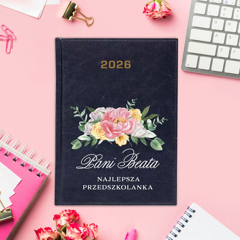 Notatnik z ciemnoniebieską okładką z kwiatowym wzorem i napisem 'Pani Beata Najlepsza Przedszkolanka', na rok 2026, leżący na różowym stole obok klawiatury, notesu i różowych kwiatów.