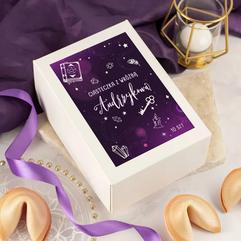 Biały kartonowy pudełko z fioletową grafiką, na której znajdują się rysunki związane z wróżbami, takie jak klucz, świece, kryształy i gwiazdy, z napisem 'Ciasteczka z wróżbą Andrzejkowa'. Obok leżą dwa ciasteczka wróżbowe i fioletowa wstążka na tle purpurowego materiału.