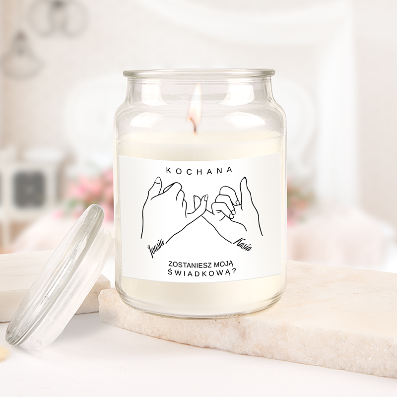 Świeca w szklanym słoiku z etykietą, na której są narysowane splecione dłonie oraz napisy 'Kochana', 'Jasia', 'Kasia' i pytanie 'Zostaniesz moją świadkową?'. Świeca zapalona, tle rozmyte z elementami dekoracyjnymi.