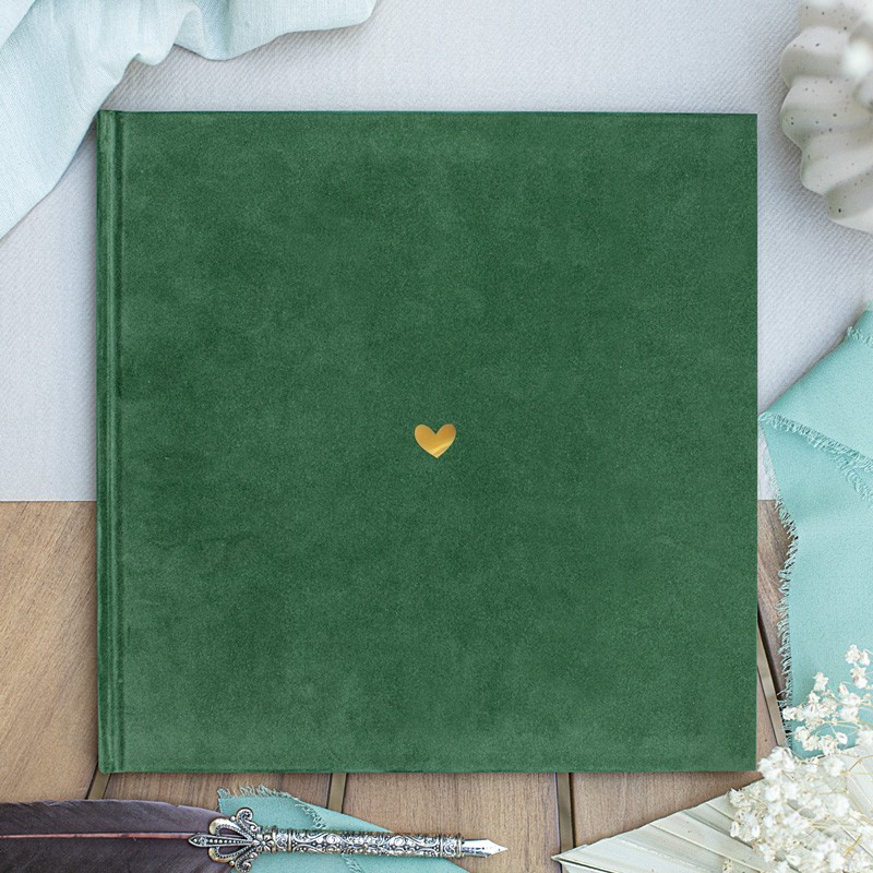 Zielony album na zdjęcia z małym, złotym serduszkiem na okładce, leżący na drewnianym stole z materiałami w tle.