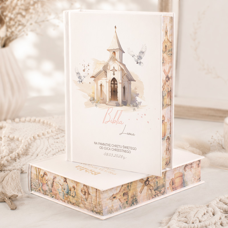 Biblia w białej okładce z ilustracją kościoła, ozdobiona pastelowymi motywami i datą chrztu.