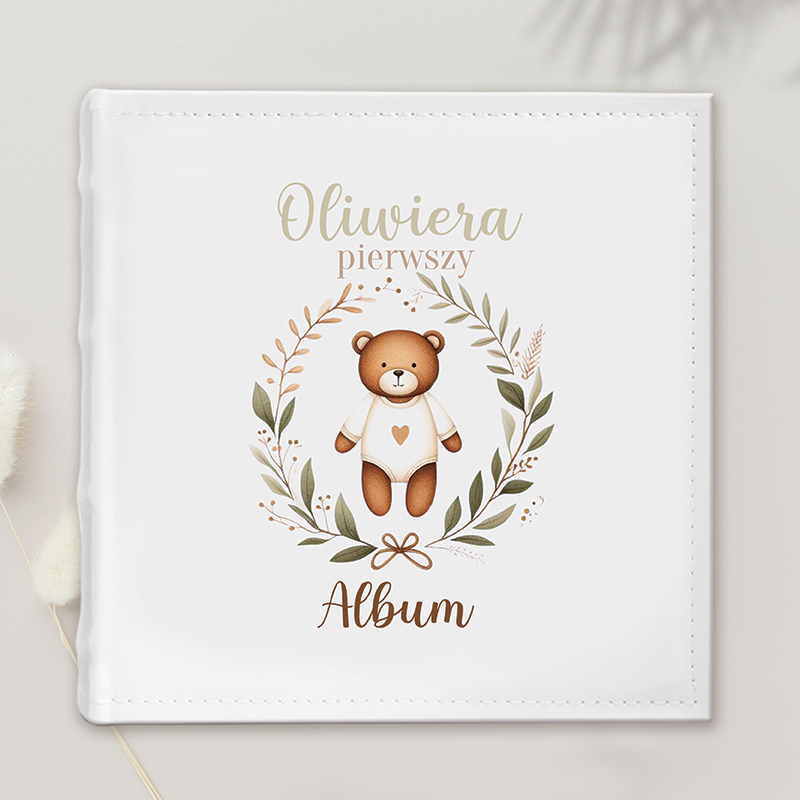 Biały album z ilustracją misia w białym ubranku, otoczonym zielonymi i brązowymi liśćmi, z napisem 'Oliwiera pierwszy Album'.