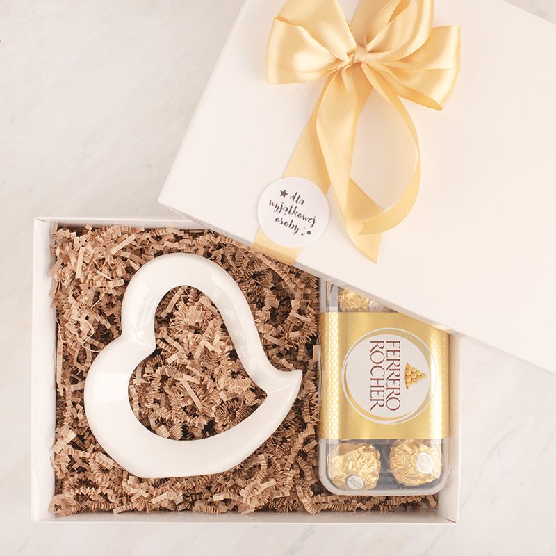Biała figurka w kształcie serca oraz pudełko z pralinkami Ferrero Rocher w eleganckim opakowaniu z złotą wstążką.