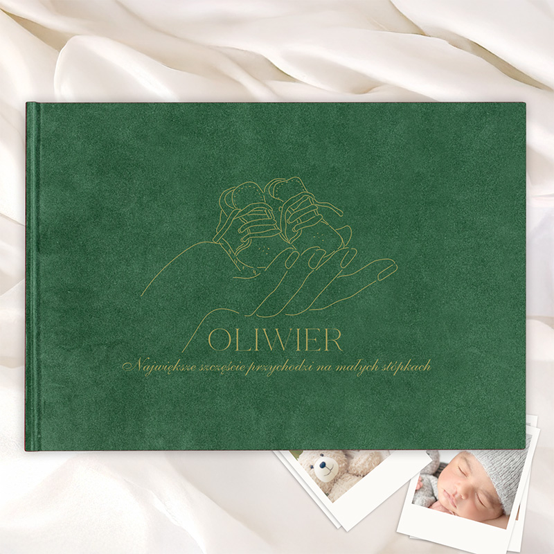 Zielona okładka książki z minimalistycznym rysunkiem dłoni trzymającej małe buciki, z napisem 'OLIWIER' i podtytułem 'Największe szczęście przychodzi na małych stópkach', położona na jasnym tle z materiału; w tle widoczne zdjęcia dziecka i pluszowego misia.