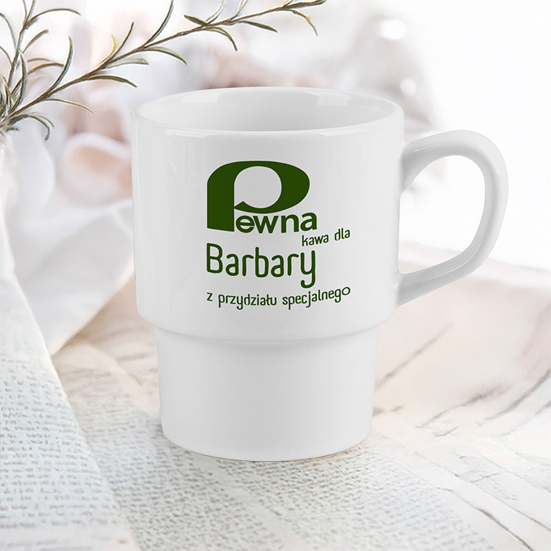 Biały kubek z zielonym nadrukiem, na którym widnieje napis 'Pewna kawa dla Barbary z przydziału specjalnego', ustawiony na tle blatu z otwartą książką i jasnym tłem.
