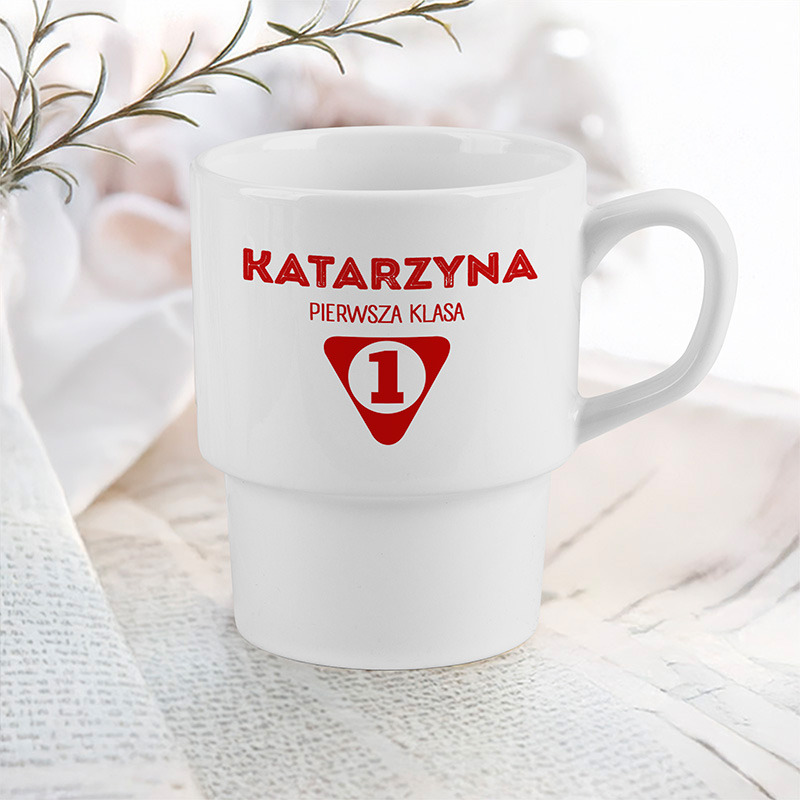 Biały kubek z czerwonym napisem "KATARZYNA PIERWSZA KLASA" oraz czerwonym symbolem oznaczającym numer 1, ustawiony na tle papieru i gałązki.