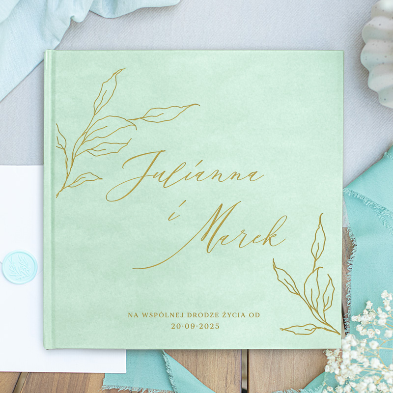 Zielona okładka książki z imionami "Julianna i Marek" napisanymi złotymi literami, zawierająca kwiatowy wzór oraz datę 20-09-2025, leżąca na tle stylowych akcesoriów.