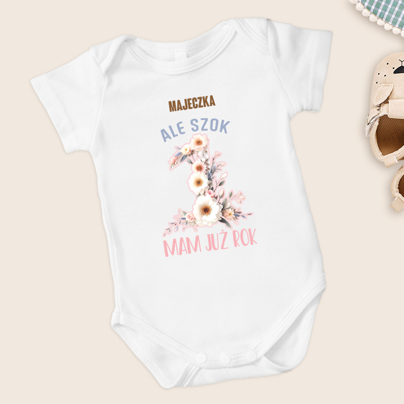 Body niemowlęce w kolorze białym z napisem 'MAJECZKA ALE SZOK 1 MAM JUŻ ROK' oraz ozdobnym kwiatowym motywem wokół liczby 1, umieszczone na jasnym tle obok bucików niemowlęcych.