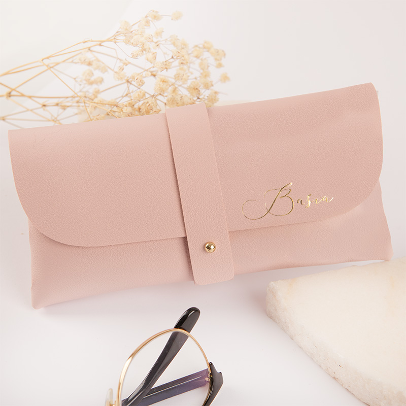 Różowa torebka w formie kopertówki z metalowym zapięciem, na której znajduje się złote imię 'Basira', obok leżą okulary przeciwsłoneczne oraz gałązka suszonych kwiatów na jasnym tle.