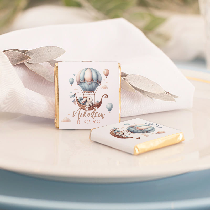 Dwa małe czekoladki na talerzu, jedna z nadrukiem przedstawiającym balon na gorące powietrze oraz napisem 'Nikodem 19 lipca 2026', obok biała serwetka ze srebrnymi dekoracjami.