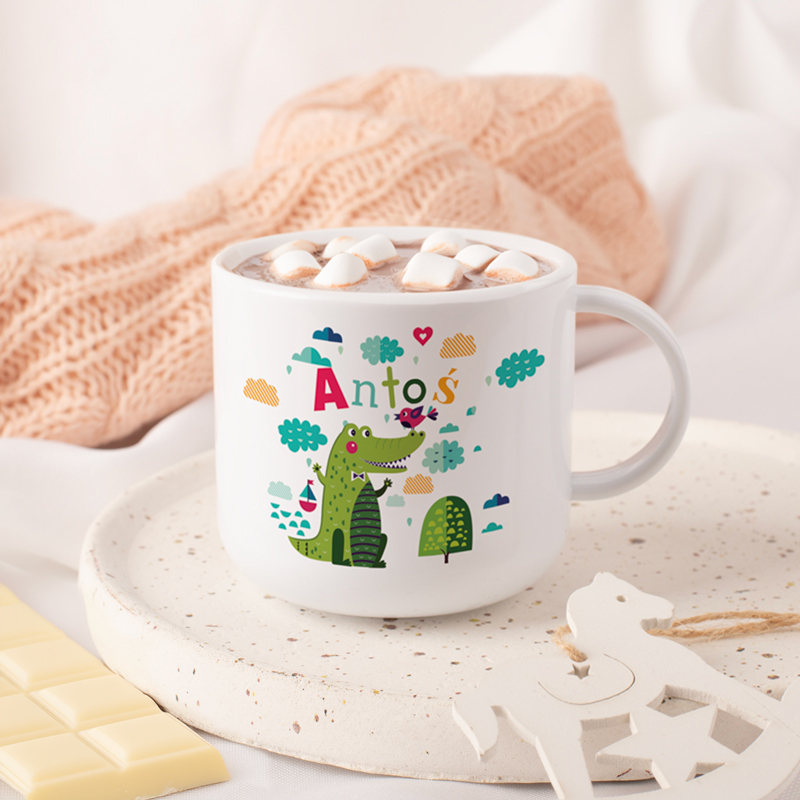 Biały kubek z czekoladą na gorąco, pokryty pianą i piankami marshmallow, zdobiony kolorowymi ilustracjami, w tym zielonym krokodylem, drzewkami i napisem 'Antos'. Kubek stoi na białym talerzu obok kawałków białej czekolady, otoczony beżowym swetrem i jasnym tłem.