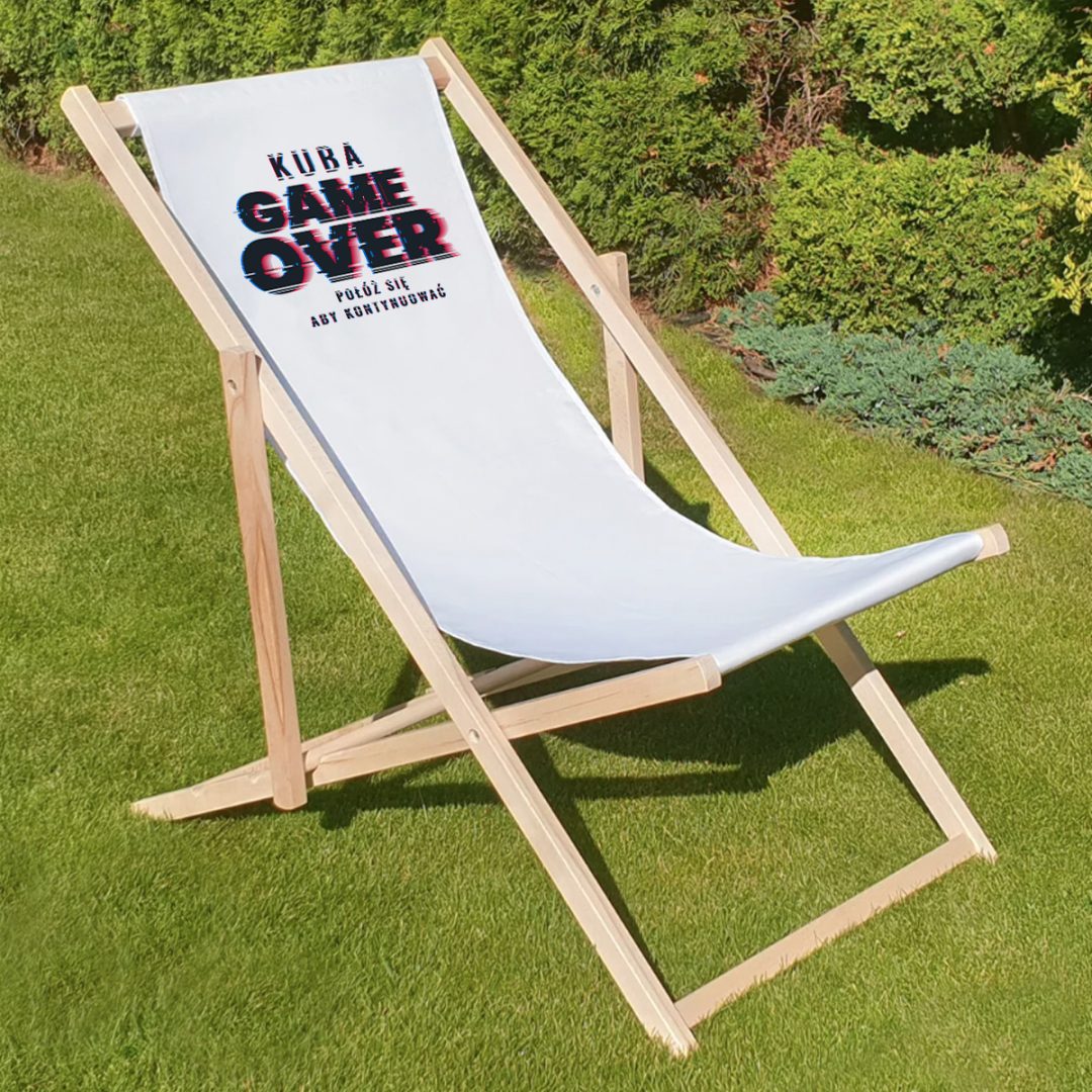 Biały leżak na trawie z napisem "KUBA GAME OVER" oraz dodatkowym tekstem pod nim, w stylu retro. Tło zielone, z niskimi krzewami w oddali.