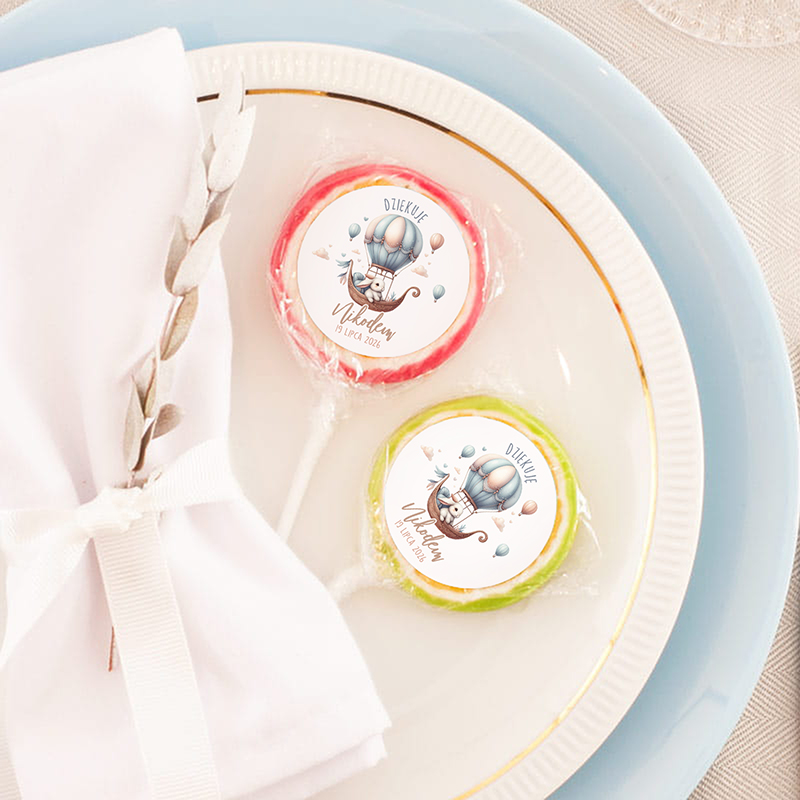 Talerz z niebieskim brzegiem, na którym znajdują się dwa lizaki: czerwony i zielony, z kolorowymi etykietami z napisem 'Dziękuję' oraz ilustracją balonów. Obok leży biała serwetka z delikatnym wzorem i ozdobną wstążką.