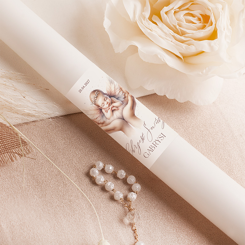 Świeca z rysunkiem aniołka i napisem 'Chrzest Święty Gabrysi' oraz datą '20.04.2023', leży na miękkim tle obok białej róży i naszyjnika.