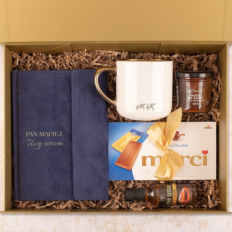 Zestaw prezentowy w złotej pudełku, zawierający niebieski notes z napisem 'PAN MACIEJ Uczy sercem', biały kubek z napisem 'MK', słoiczek z miodem z przyprawami, tabliczkę czekolady marki 'merci' z żółtą wstążką, oraz butelkę syropu karmelowego na brązowym tle.