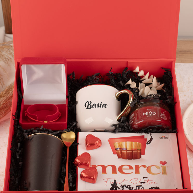 Zestaw prezentowy w czerwonym pudełku zawierający kubek z imieniem 'Basia', pierścionek w eleganckim opakowaniu, złotą łyżeczkę, słoik z miodem, zestaw czekoladek 'Merci' i dekoracyjne elementy, wszystkie umieszczone na czarnym wypełnieniu.