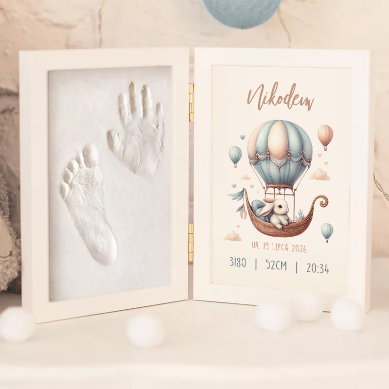 Otwarta ramka z odciskami rączki i nóżki dziecka po lewej stronie oraz ilustrowanym kartonem po prawej, zawierającym imię 'Nikodem', datę urodzenia, wagę i wzrost dziecka, wszystko ozdobione balonami na gorące powietrze.