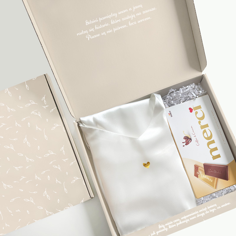 Otwarta pudełko prezentowe z białą satynową apaszką z małym złotym serduszkiem oraz czekoladkami marki 'merci' na tle jasnego wnętrza z delikatnym wpisem na pokrywie.