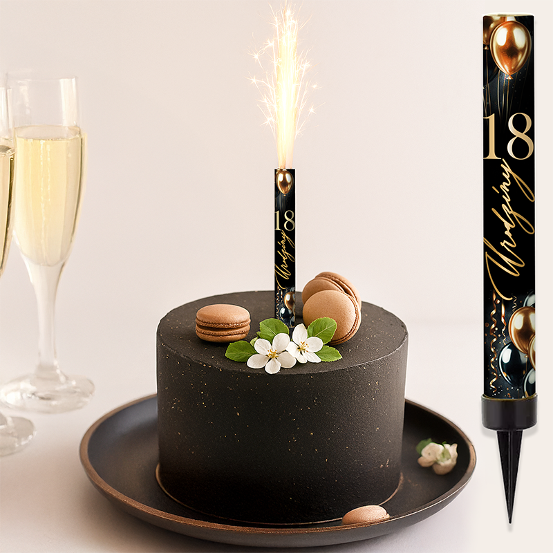 Czarny tort urodzinowy z fajerwerkami w kształcie świeczki, ozdobiony białymi kwiatami i makaronikami, obok dwa kieliszki z szampanem na tle jasnej ściany.
