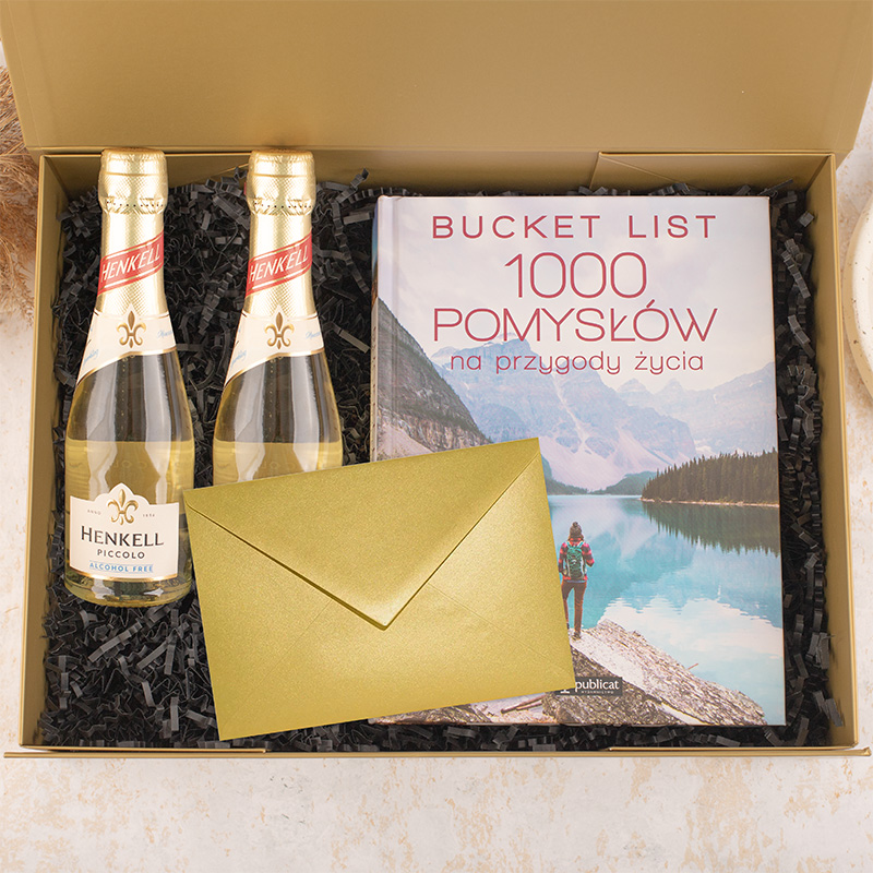 Zestaw prezentowy w złotym pudełku zawierający dwie butelki bezalkoholowego szampana Henkell, książkę zatytułowaną 'Bucket List 1000 pomysłów na przygody życia' oraz złotą kopertę, ułożone na czarnych wiórkach papierowych.