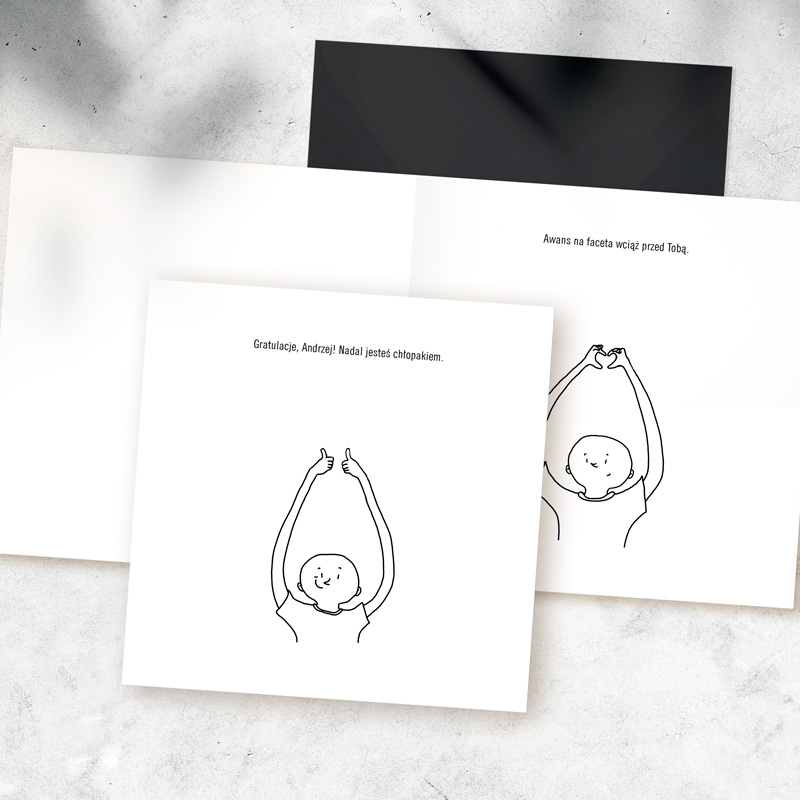 Rysunek przedstawiający postać z podniesionymi rękami i unoszącymi kciukami, z uśmiechniętą twarzą. W tle widoczne są strony kartki z napisem: 'Gratulacje, Andrzej! Nadal jesteś chłopakiem.' oraz 'Awans na faceta wciąż przed Tobą.'