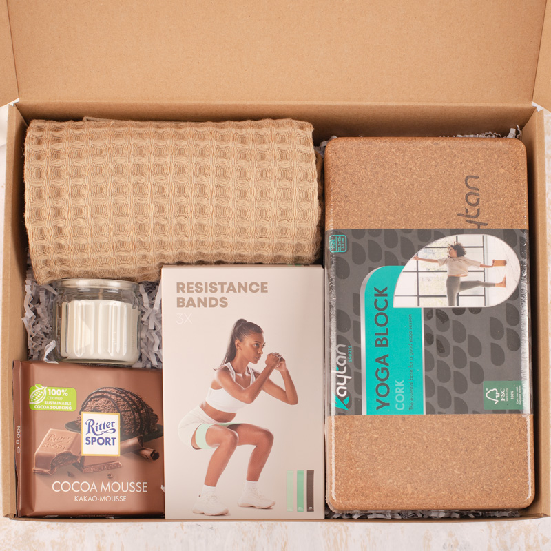 Zestaw zawierający: bezbarwny wysoki ręcznik, zestaw trzech gum oporowych, świecę w szklanym słoiku, tabliczkę czekolady Ritter Sport o smaku musowym oraz blok do jogi z korka, wszystko w kartonowym pudełku wypełnionym papierem wypełniającym.