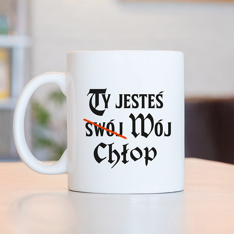 Biały kubek z czarnym napisem "Ty jesteś swoj Chłop". Część słowa "Wój" jest przekreślona na rudo, co sugeruje zmianę w treści.