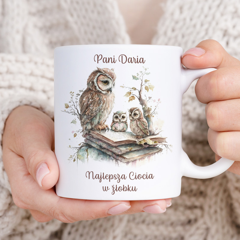 Kubek z obrazkiem sów siedzących na otwartej książce, z napisem 'Pani Daria' i 'Najlepsza Ciocia w żłobku', trzymany w dłoniach osoby, w tle widoczny jasny sweter.