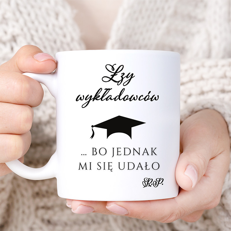 Biała filiżanka trzymana przez osobę, z napisem 'Łzy wykładowców ... BO JEDNAK MI SIĘ UDAŁO' oraz grafiką czapki absolwenta.