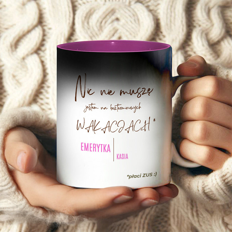 Kubek z napisem 'Nic nie muszę, jestem na bezterminowych WAKACJACH* EMERYTKA KASIA', trzymany w dłoniach osoby w beżowym swetrze. Wnętrze kubka jest różowe.