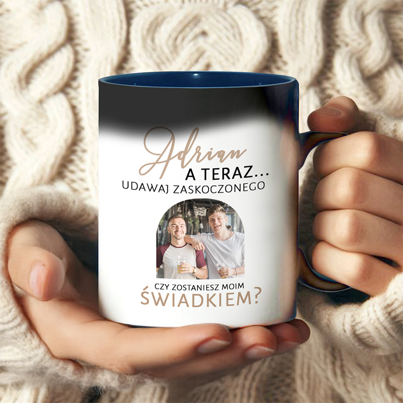 Kubek w dłoniach, z napisem 'Adrian, a teraz... udawaj zaskoczonego' oraz pytaniem 'Czy zostaniesz moim świadkiem?', na tle zdjęcia dwóch uśmiechających się mężczyzn trzymających szklanki z napojem, na tle z przędzy swetra.